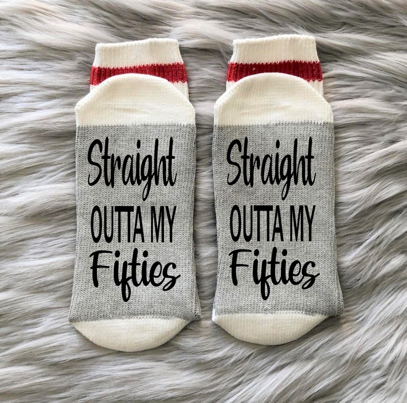 Puede incluir: Un par de calcetines blancos hasta el tobillo con una franja roja en la parte superior. Los calcetines tienen una franja gris en el medio con el texto "Straight outta my fifties" impreso en negro.