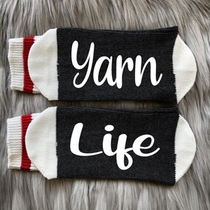 Puede incluir: Un par de calcetines negros con las palabras "Yarn" y "Life" en escritura blanca. Los calcetines tienen punteras blancas y pu&ntilde;os a rayas rojas y blancas. Los calcetines se exhiben sobre una superficie gris y peluda.