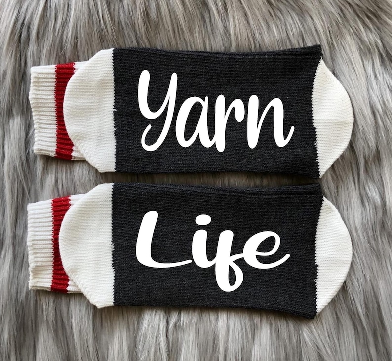 Puede incluir: Un par de calcetines negros con las palabras "Yarn" y "Life" en escritura blanca. Los calcetines tienen punteras blancas y pu&ntilde;os a rayas rojas y blancas. Los calcetines se exhiben sobre una superficie gris y peluda.