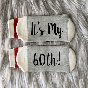 Puede incluir: Un par de calcetines grises con rayas rojas y blancas. Los calcetines tienen el texto "It's My 60th!" impreso en ellos.