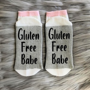 May include: A pair of gray and white socks with a pink stripe at the top. The socks have the text "Gluten Free Babe" printed on them in black.