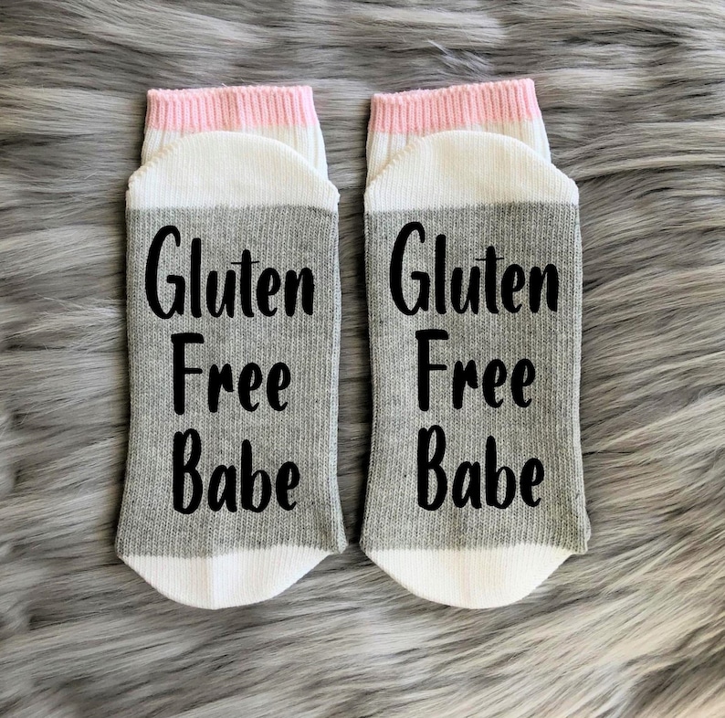 May include: A pair of gray and white socks with a pink stripe at the top. The socks have the text "Gluten Free Babe" printed on them in black.