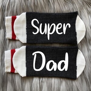 May include: A pair of black socks with white lettering. The socks have red and white stripes at the top. The text on the socks reads "Super Dad".