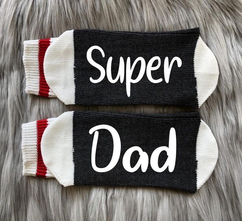 May include: A pair of black socks with white lettering. The socks have red and white stripes at the top. The text on the socks reads "Super Dad".