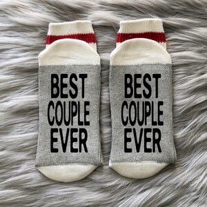 Puede incluir: Un par de calcetines grises y blancos con rayas rojas. Los calcetines tienen el texto "Best Couple Ever" impreso en negro.