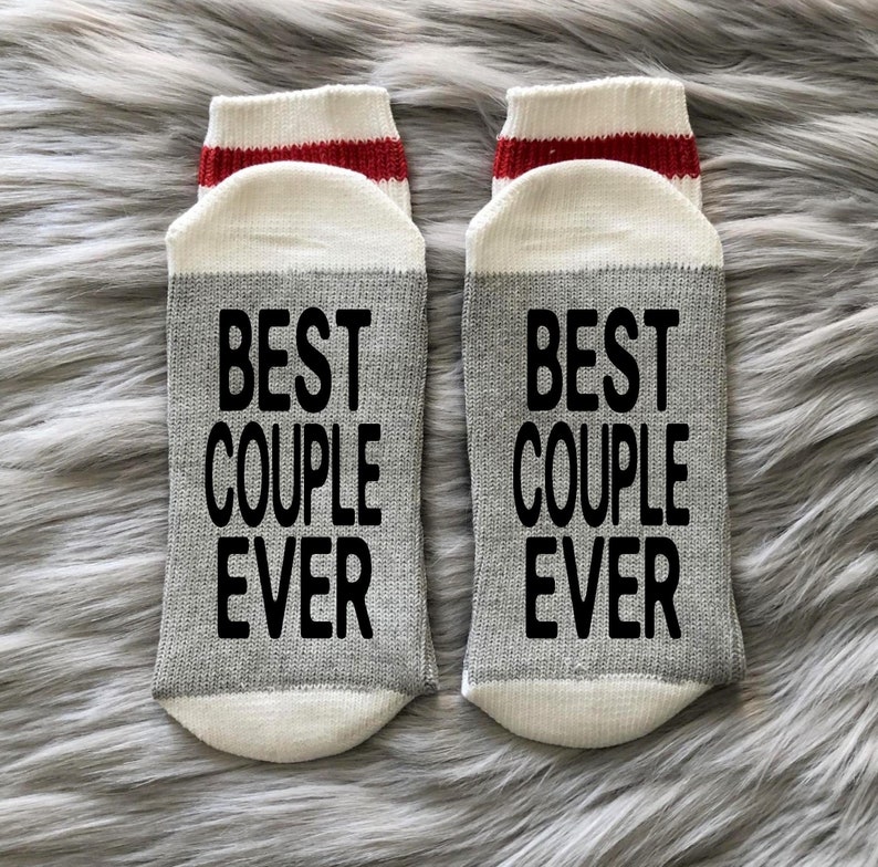 Puede incluir: Un par de calcetines grises y blancos con rayas rojas. Los calcetines tienen el texto "Best Couple Ever" impreso en negro.