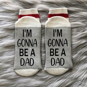 Op de afbeelding: Een paar grijze en witte sokken met rode strepen aan de bovenkant. De sokken hebben de tekst "I'm gonna be a dad" in zwart erop gedrukt.