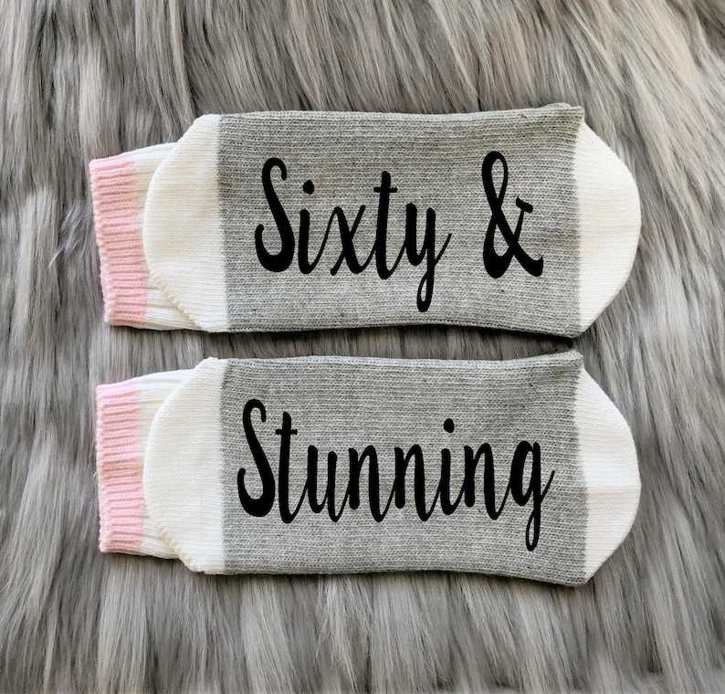 Puede incluir: Un par de calcetines grises y blancos con ribete rosa. Los calcetines tienen el texto "Sixty & Stunning" impreso en negro.