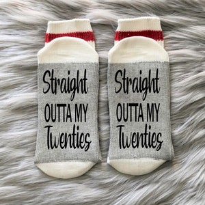 May include: A pair of gray and white ankle socks with a red stripe at the top. The socks have the text "Straight outta my twenties" printed on them.