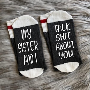 May include: A pair of dark gray socks with white toes and cuffs. One sock reads "MY SISTER AND I" in white script, and the other reads "TALK SHIT ABOUT YOU". The cuffs have a red stripe.