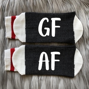 K&ouml;nnte beinhalten: Ein Paar schwarze Socken mit wei&szlig;en Buchstaben. Eine Socke sagt "GF" und die andere sagt "AF". Die Socken haben einen wei&szlig;en Bund mit einem roten Streifen.
