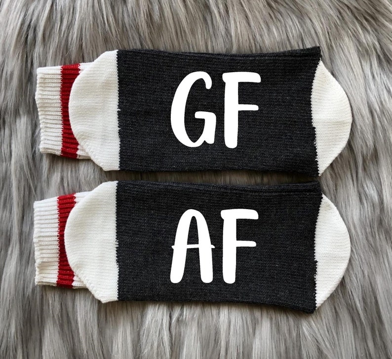 K&ouml;nnte beinhalten: Ein Paar schwarze Socken mit wei&szlig;en Buchstaben. Eine Socke sagt "GF" und die andere sagt "AF". Die Socken haben einen wei&szlig;en Bund mit einem roten Streifen.