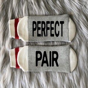 Puede incluir: Un par de calcetines grises con pu&ntilde;os blancos y rayas rojas. Un calcet&iacute;n dice "PERFECT" y el otro dice "PAIR".