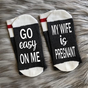 Op de afbeelding: Een paar zwarte sokken met witte bies. Op de ene sok staat "GO easy ON ME" en op de andere staat "MY WIFE is PREGNANT".