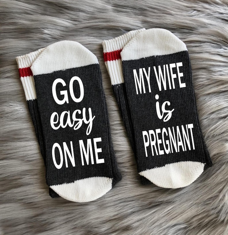 Op de afbeelding: Een paar zwarte sokken met witte bies. Op de ene sok staat "GO easy ON ME" en op de andere staat "MY WIFE is PREGNANT".