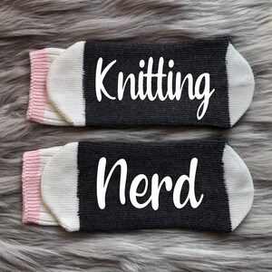 Puede incluir: Dos pares de calcetines grises con texto blanco "Knitting Nerd". Los calcetines tienen punteras blancas, pu&ntilde;os rosas y se muestran sobre una superficie gris y difusa. Para tejedores.