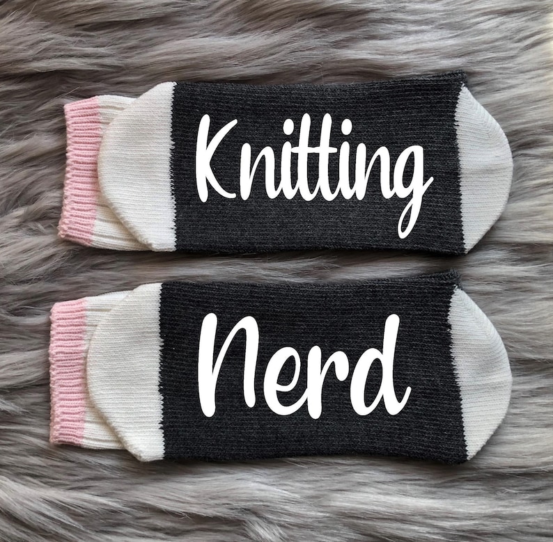 Puede incluir: Dos pares de calcetines grises con texto blanco "Knitting Nerd". Los calcetines tienen punteras blancas, pu&ntilde;os rosas y se muestran sobre una superficie gris y difusa. Para tejedores.