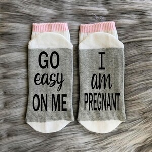 Op de afbeelding: Een paar grijze en witte sokken met roze bies. E&eacute;n sok zegt "GO easy ON ME" en de andere zegt "I am PREGNANT".