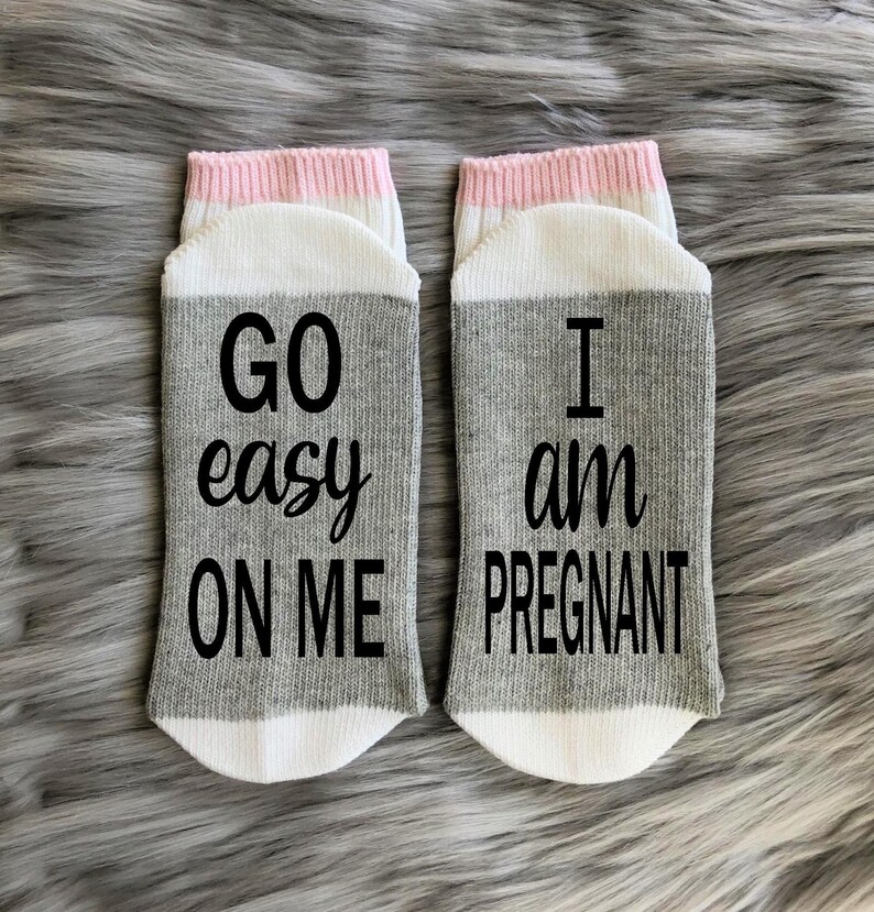 Op de afbeelding: Een paar grijze en witte sokken met roze bies. E&eacute;n sok zegt "GO easy ON ME" en de andere zegt "I am PREGNANT".