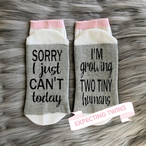 May include: A pair of gray and white socks with pink trim. One sock says "Sorry I just can't today" and the other says "I'm growing two tiny humans". The socks are labeled "Expecting Twins".