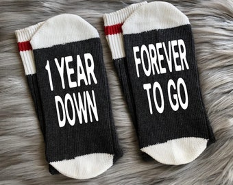 May include: A pair of black socks with white trim and red stripes. One sock says "1 year down" and the other says "forever to go".