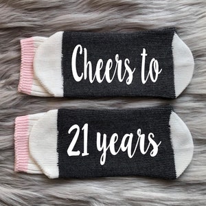 May include: A pair of black and white socks with pink trim. The socks have the text "Cheers to 21 years" written in white.