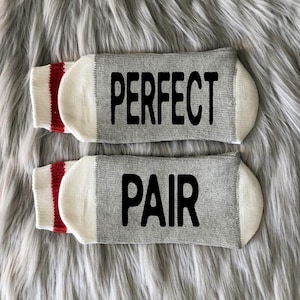 Puede incluir: Un par de calcetines grises con pu&ntilde;os blancos y rayas rojas. Un calcet&iacute;n dice "PERFECT" y el otro dice "PAIR" en letras negras.
