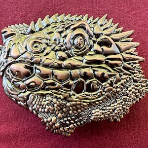 May include: A detailed, silver-toned metal belt buckle with a textured, reptilian design. The buckle features a close-up of a lizard's face with a scaly texture.