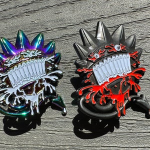 May include: Two enamel pins featuring a cartoon monster design. The pins are black and iridescent with white teeth and red and white dripping paint.