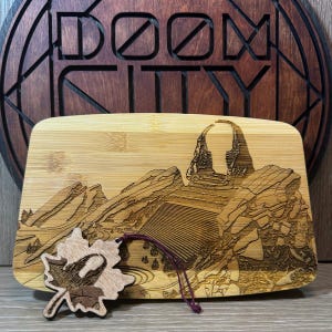 Puede incluir: Tabla de cortar rectangular de bambú con un diseño de paisaje grabado con láser. El diseño presenta una cordillera y una masa de agua. Un adorno de madera en forma de hoja está sujeto con una cuerda morada. La palabra "DOOM CITY" está grabada en un fondo de madera.
