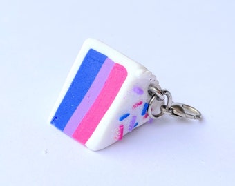 Puede incluir: Un pequeño amuleto de pastel blanco con glaseado azul, morado y rosa y chispas de colores. El amuleto está unido a un llavero plateado.