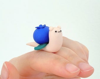 May include: A small, white snail figurine with a blue blueberry on its back. The snail has a cute, cartoon-like face with black eyes and a small mouth. The blueberry is a bright blue color and has a green leaf on top.