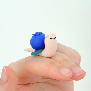 May include: A small, white snail figurine with a blue blueberry on its back. The snail has a cute, cartoon-like face with black eyes and a small mouth. The blueberry is a bright blue color and has a green leaf on top.