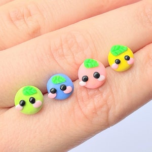 May include: Four small, colorful clay charms with cute faces and green leaves. Each charm is a different color: green, blue, pink, and yellow.