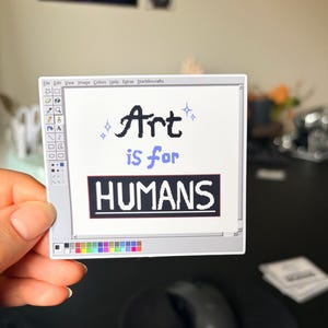 May include: A white sticker with a computer-like interface design. The sticker features the text "Art is for HUMANS" in black and blue. The sticker is held by a hand, with a black mouse and mousepad in the background.