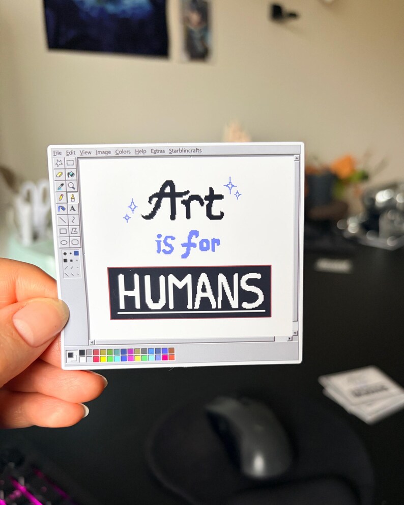 May include: A white sticker with a computer-like interface design. The sticker features the text "Art is for HUMANS" in black and blue. The sticker is held by a hand, with a black mouse and mousepad in the background.