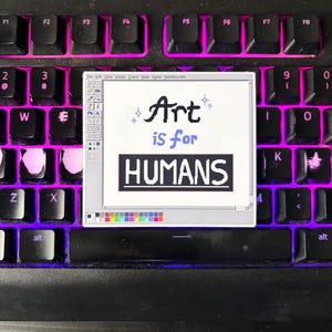 May include: A sticker featuring a retro computer-style design with the words "Art is for HUMANS." The sticker is placed on a black keyboard with pink backlighting. The design mimics an old computer program interface.