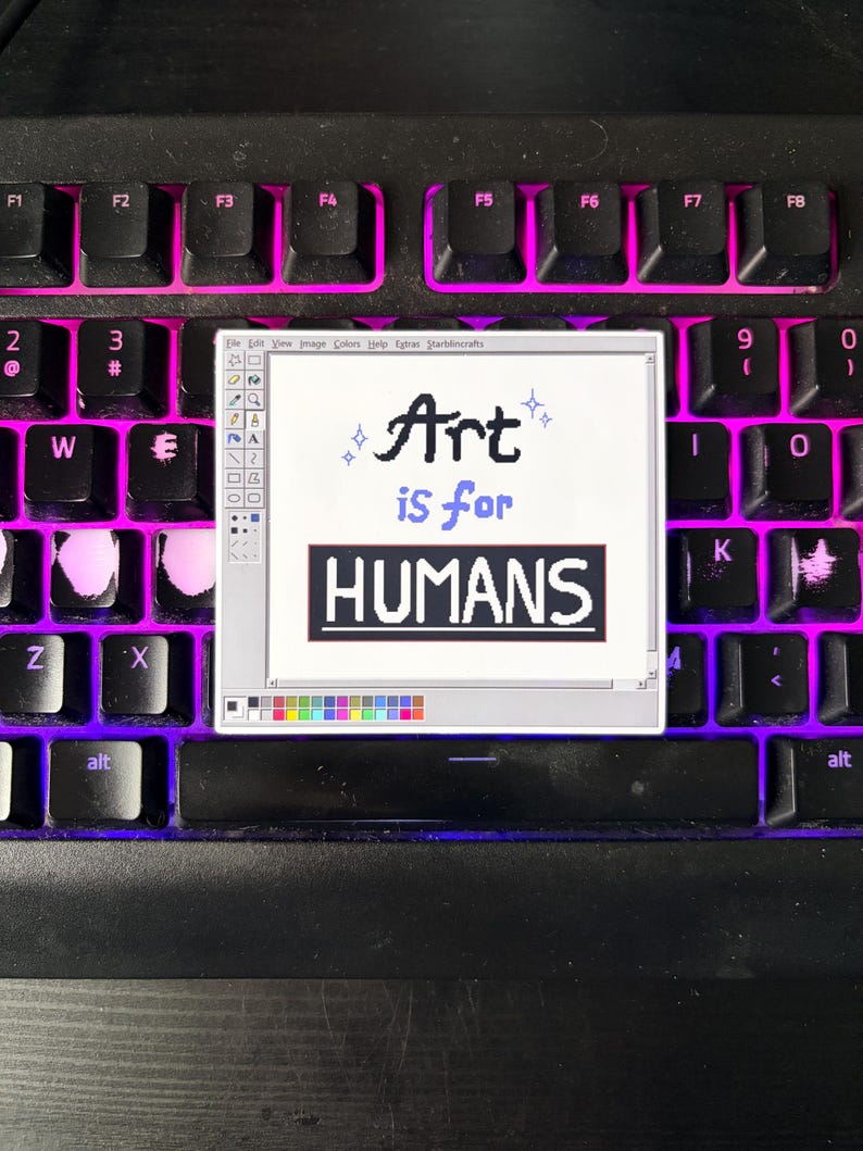 May include: A sticker featuring a retro computer-style design with the words "Art is for HUMANS." The sticker is placed on a black keyboard with pink backlighting. The design mimics an old computer program interface.