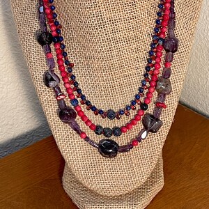 Op de afbeelding: Een meerrijige ketting met rode, paarse en blauwe kralen. De ketting is gemaakt van verschillende edelstenen en wordt getoond op een jutepop.