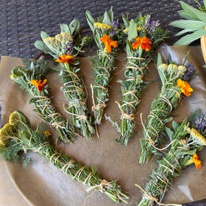 Könnte beinhalten: Mehrere Bündel getrockneter Kräuter und Blumen, darunter Ringelblumen, Salbei und Lavendel, sind mit Bindfaden gebunden. Die Bündel sind auf einer braunen Oberfläche angeordnet und zeigen die natürlichen Farben und Texturen. Diese aromatischen Bündel werden wahrscheinlich zum Räuchern oder für die Aromatherapie verwendet.