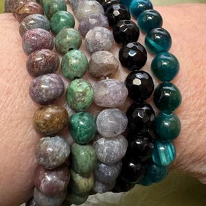 May include: Five beaded bracelets stacked on a wrist. The bracelets are made of different colored stones, including purple, green, black, and blue.