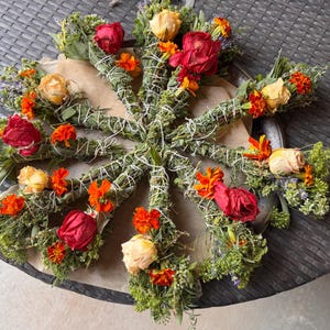 May include: A circular arrangement of eight dried herb bundles tied with white string. Each bundle features a mix of green foliage, dried red and yellow roses, and small orange marigold flowers, creating a vibrant, multi-colored floral display.