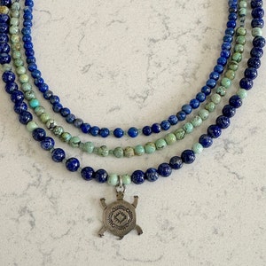 Può includere: Una collana a più fili con perline di pietre preziose blu e verdi e un ciondolo a forma di tartaruga in argento.