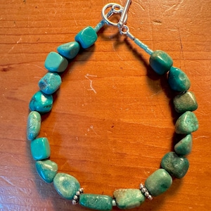 Peut inclure: Bracelet de perles en turquoise avec fermoir à bascule en argent. Le bracelet est composé de pierres de turquoise polies et irrégulières dans des tons de vert et de bleu. Le fermoir et les petites perles d'argent ajoutent une touche d'élégance.