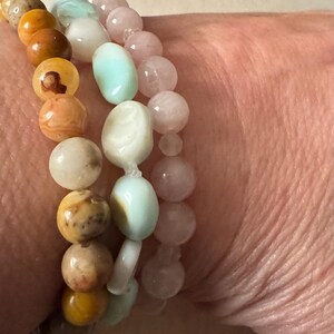 May include: Three bracelets made of natural stones. The top bracelet is made of light pink stones, the middle bracelet is made of light blue and white stones, and the bottom bracelet is made of brown and orange stones.