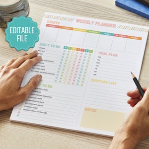 Może przedstawiać: Notatnik tygodniowy z kolorowym paskowanym wzorem. Notatnik zawiera sekcje na wydarzenia, codzienne zadania, plan posiłków, listę zakupów i notatki. Notatnik leży na drewnianej powierzchni, a ręka trzyma ołówek.