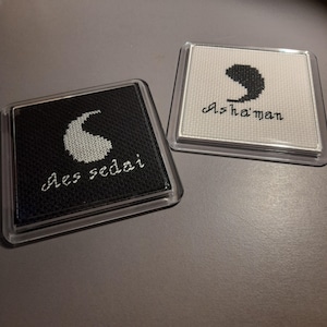 May include: Two square coasters with clear acrylic frames. One coaster has a black background with a white embroidered teardrop symbol and the text Aes sedai. The other has a white background with a black embroidered teardrop and the text Asha'man.