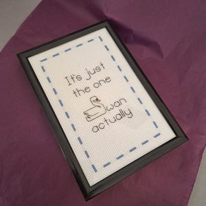 Może przedstawiać: Oprawiona praca haftu krzyżykowego z napisem "It's just the one swan actually" i ilustracją łabędzia. Dzieło sztuki jest w czarnej ramie i ma niebieskie przeszycia na białym tle. Tło to fioletowa powierzchnia.