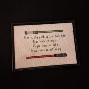 Peut inclure: Broderie au point de croix encadrée en noir. L'œuvre présente des sabres laser verts et rouges et la citation : "La peur est le chemin vers le côté obscur. La peur mène à la colère. La colère mène à la haine. La haine mène à la souffrance."