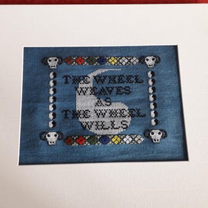 May include: A cross-stitch embroidery on blue fabric featuring the text "THE WHEEL WEAVES AS THE WHEEL WILLS" in black, centered over a white crescent shape. The border includes four ram skulls, vertical yin-yang symbols, and colorful geometric patterns.